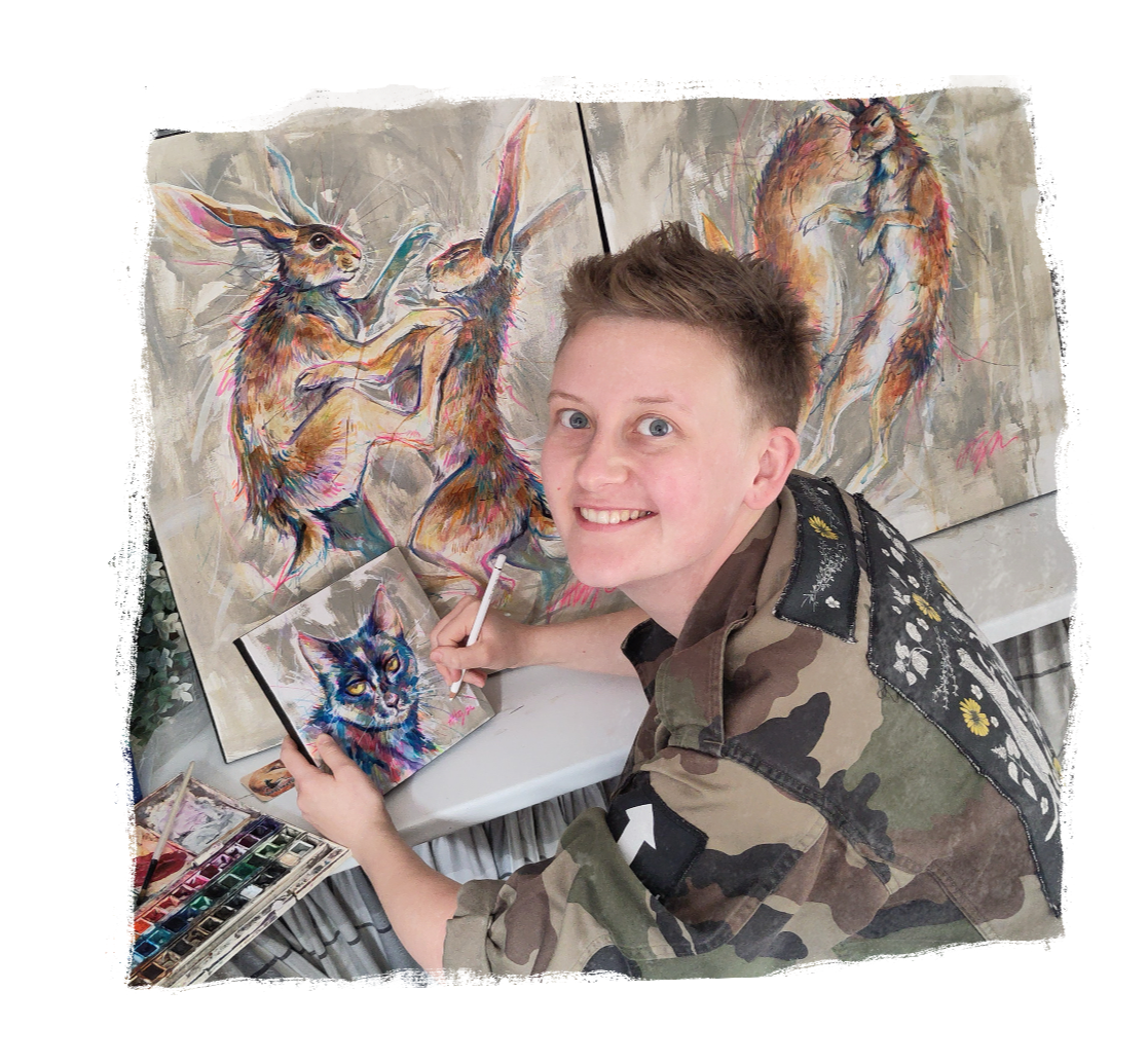 A photograph of Hoggy working on a painting. Hoggy has pale skin and dark blond hair styled into a short quiff, he's wearing a camo jacket and is sat turned towards the camera, behind are some of his paintings.